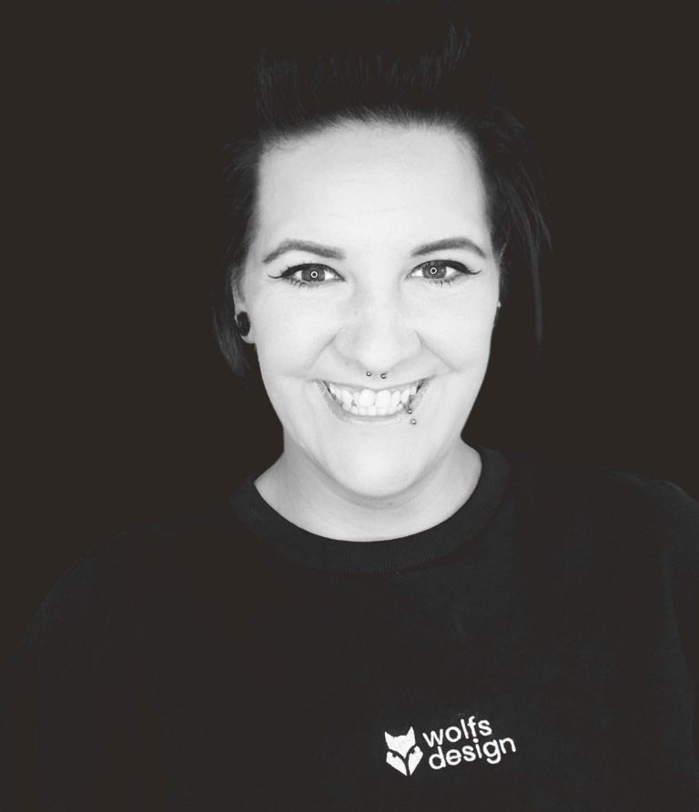 Foto Nadine Wolf Geschäftsführung wolfsdesign Eine lächelnde Person mit dunklem Shirt und dunklen Haaren vor einem schwarzen Hintergrund.